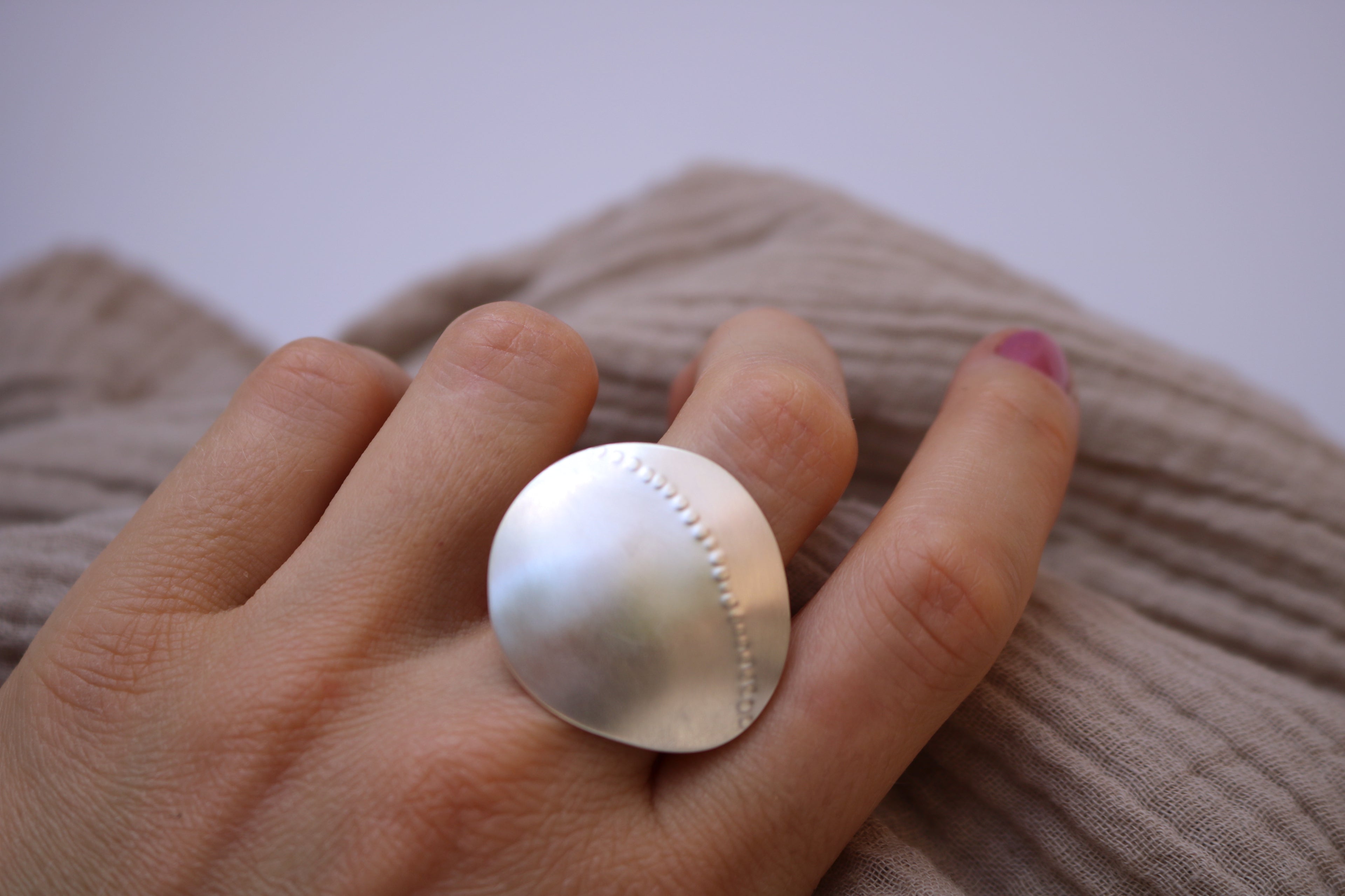 Hand wearing a large statement silver ring with a textured surface against a neutral background